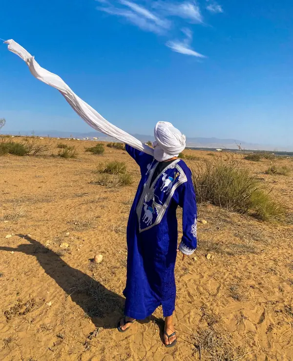 Someone in a blue robe letting a long white scarf fly in the wind on open, sandy scrubland